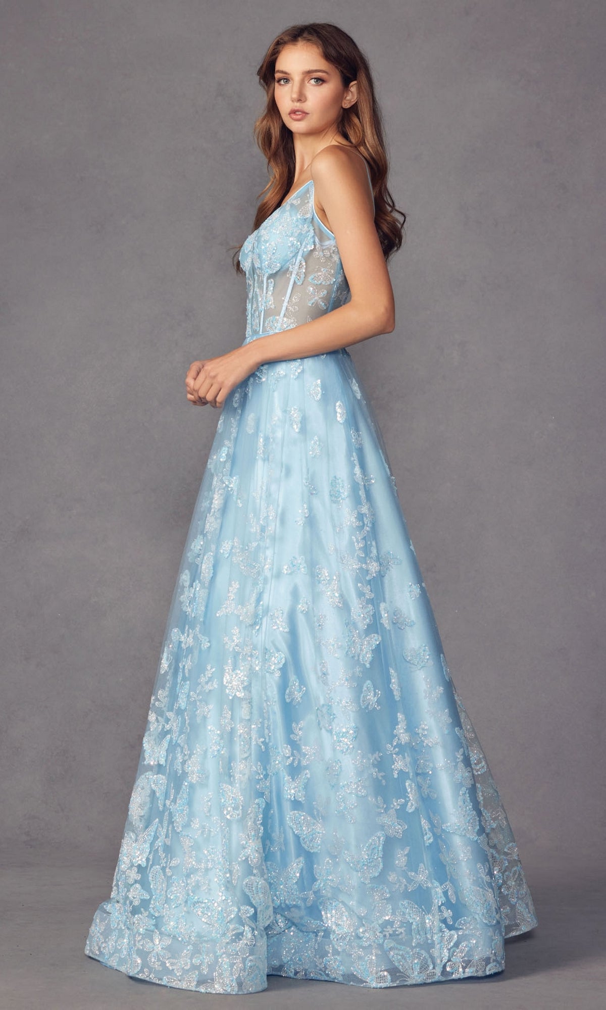 Woman wearing the Sheer-Corset Glitter-Print Long Prom Dress 2413, a sleeveless, light blue A-line gown with floral lace details, standing sideways against a plain gray background.